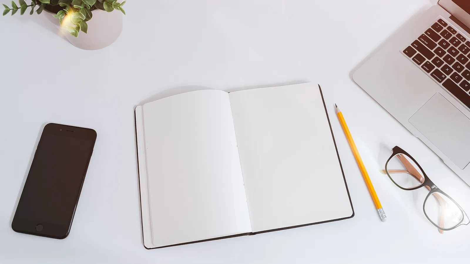 White desk with an open notebook, smartphone, glasses and laptop